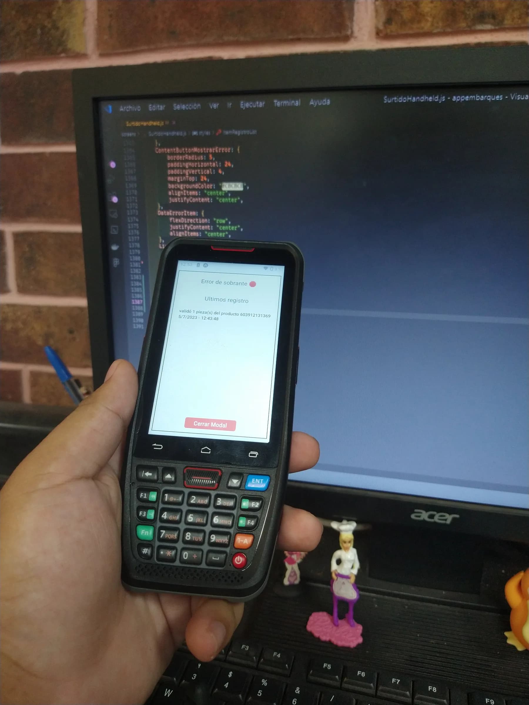 Un handheld de bodega con teclado físico en la mano, mostrando en pantalla un aviso de error de sobrante y el último registro escaneado; al fondo, el código de la app en un monitor